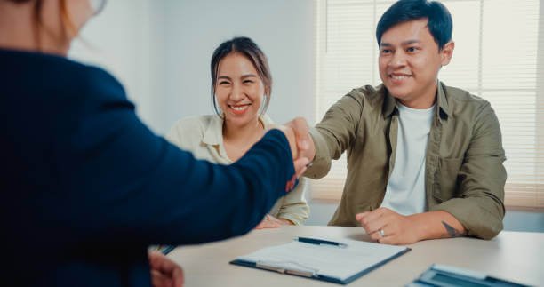 Saleswoman give house key to young married couple with casual sitting at desk feeling happy with sign peper new home contract agreement at estate office.